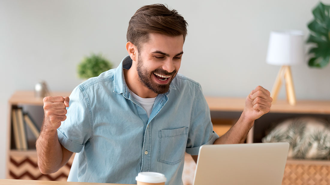Hombre sentado frente a laptop sonriendo y levantando los puños en forma de celebración