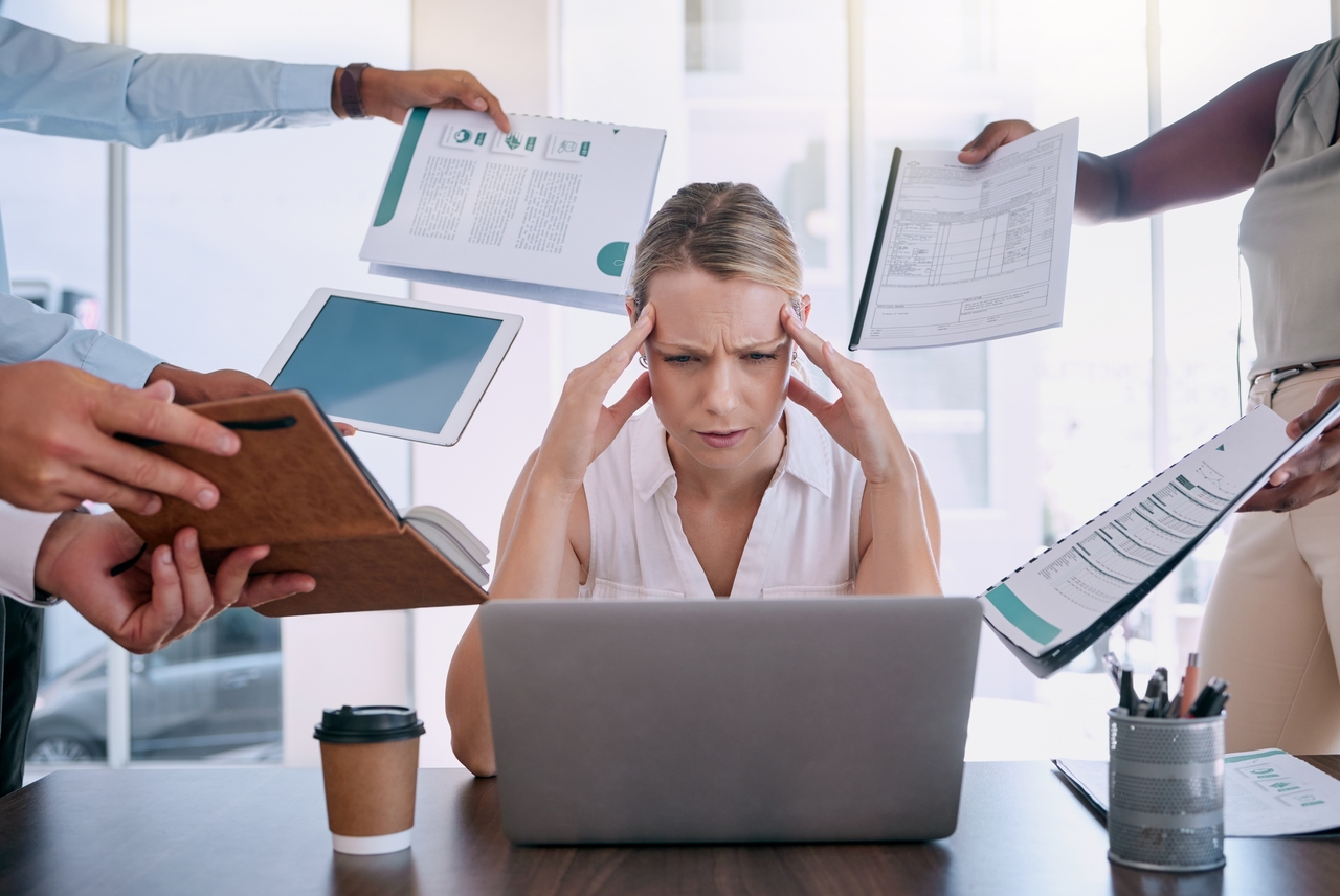 Mujer sentada frente a laptop tocándose la cabeza rodeada de personas sosteniendo cuadernillos de trabajo