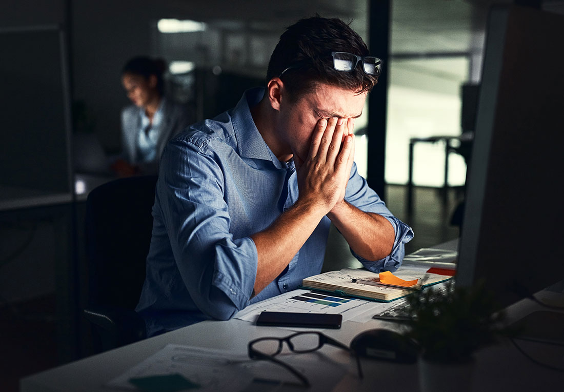 Hombre tapándose el rostro con las manos sentado frente a una computadora 