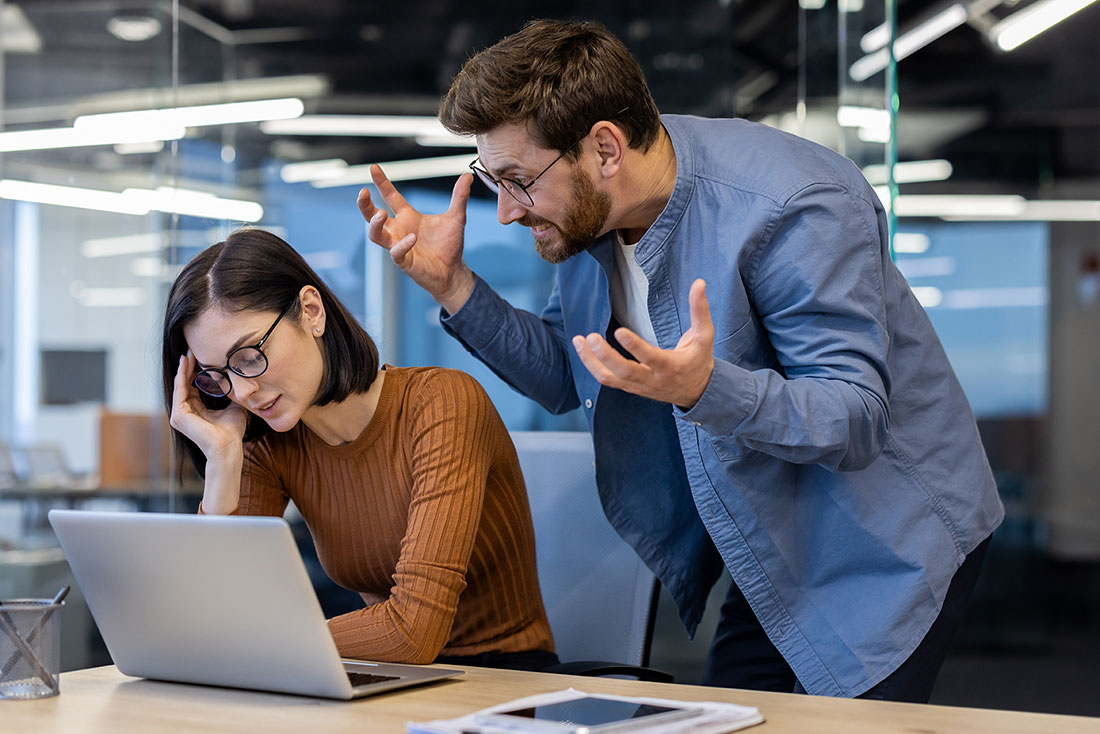 Hombre irritado en oficina hablando con una compañera de trabajo sentada frente a una computadora 