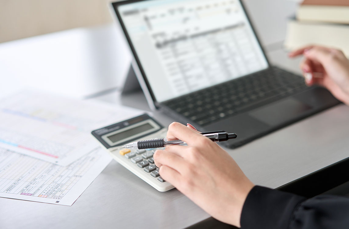 Una persona usa una calculadora mientras revisa una lista de nómina en una computadora portátil. 