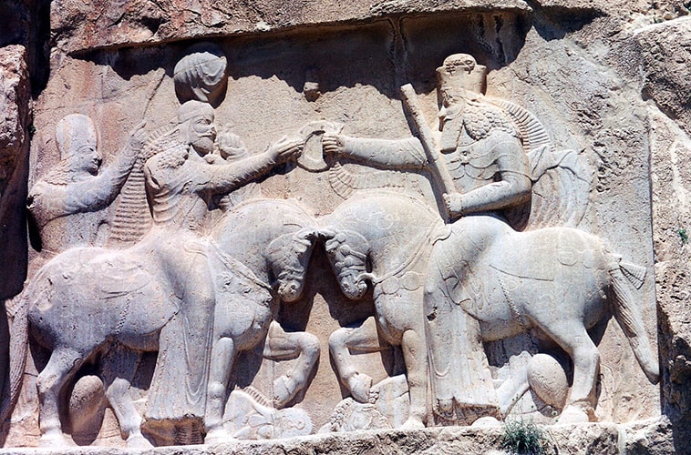 En este relieve, se muestra a dos personajes nobles que se dan muestra de cortesía o saludo, llevando en la mano izquierda el haz adivinatorio.