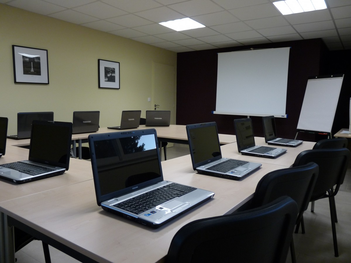 Salón con mesas dispuestas en forma rectangular, con computadoras frente a cada silla y, al fondo, un pizarrón y una pantalla.