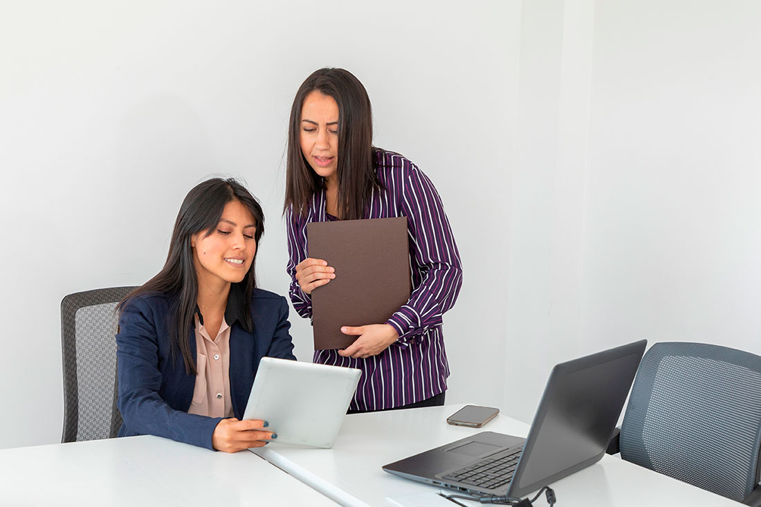 Dos mujeres conversando en una oficina frente a una Tablet y una laptop