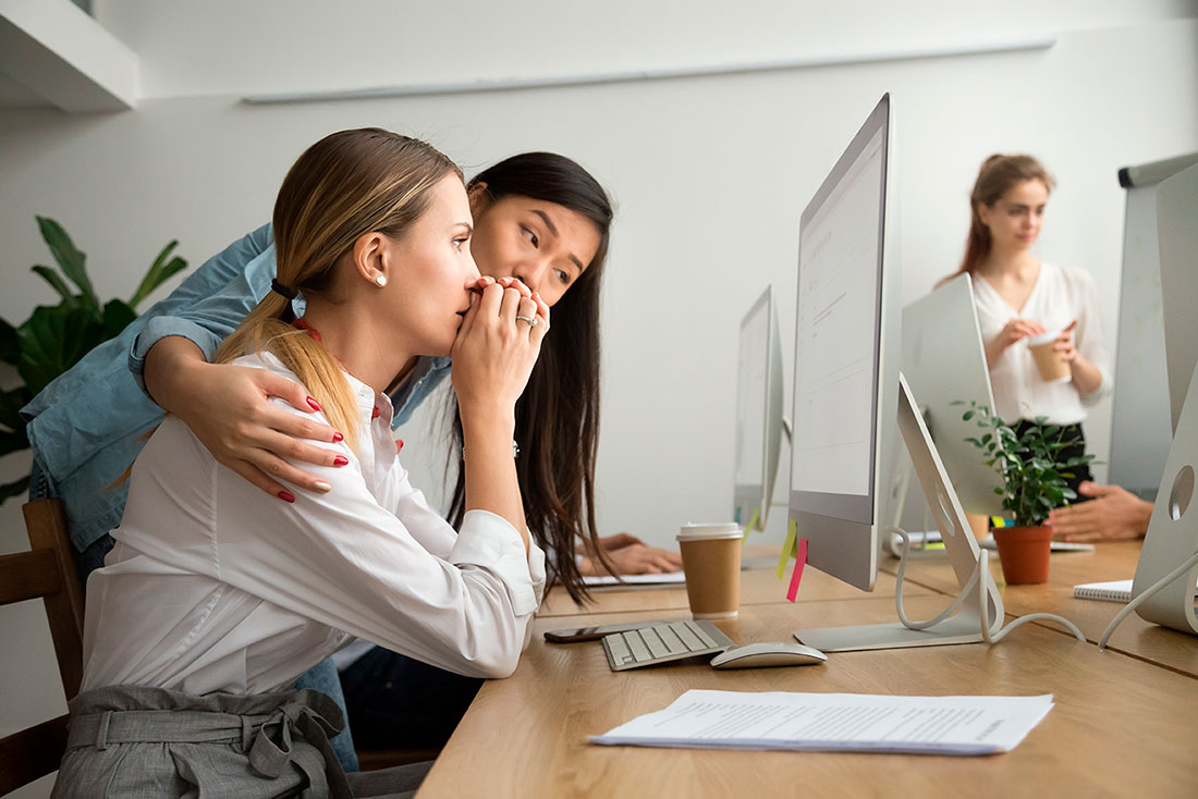 Mujer abrazando a una compañera del trabajo sentada frente a una computadora en oficina