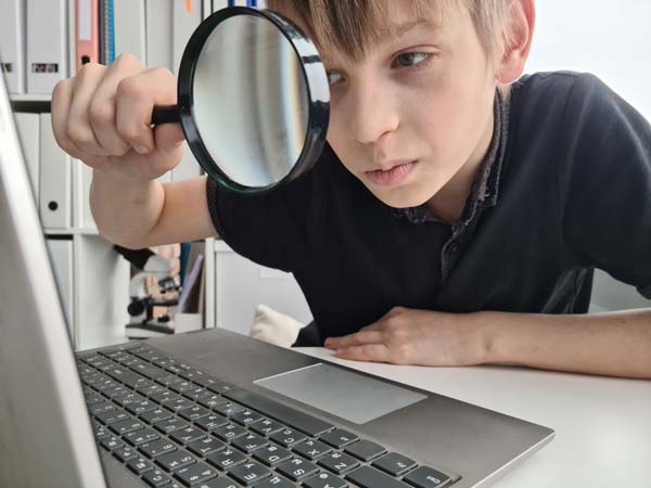 Niño observando computadora Niño con una lupa en la mano observando a través de ella una laptop