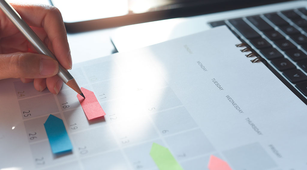 Planificación de agenda Primer plano de una mano sosteniendo un lápiz y señalando una fecha en un calendario en un escritorio junto a un teclado de computadora